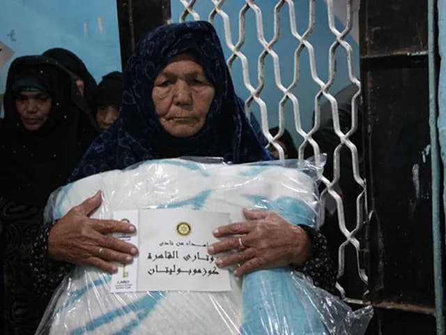 An elderly woman holding a wrapped winter blanket labelled in Arabic as a gift from the club